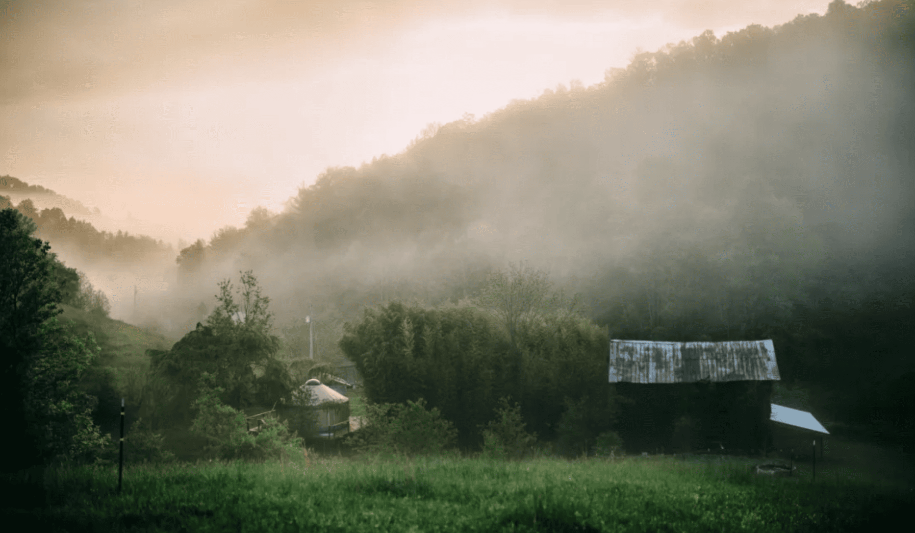A small yurt sits at the foot of a mountain on a foggy morning